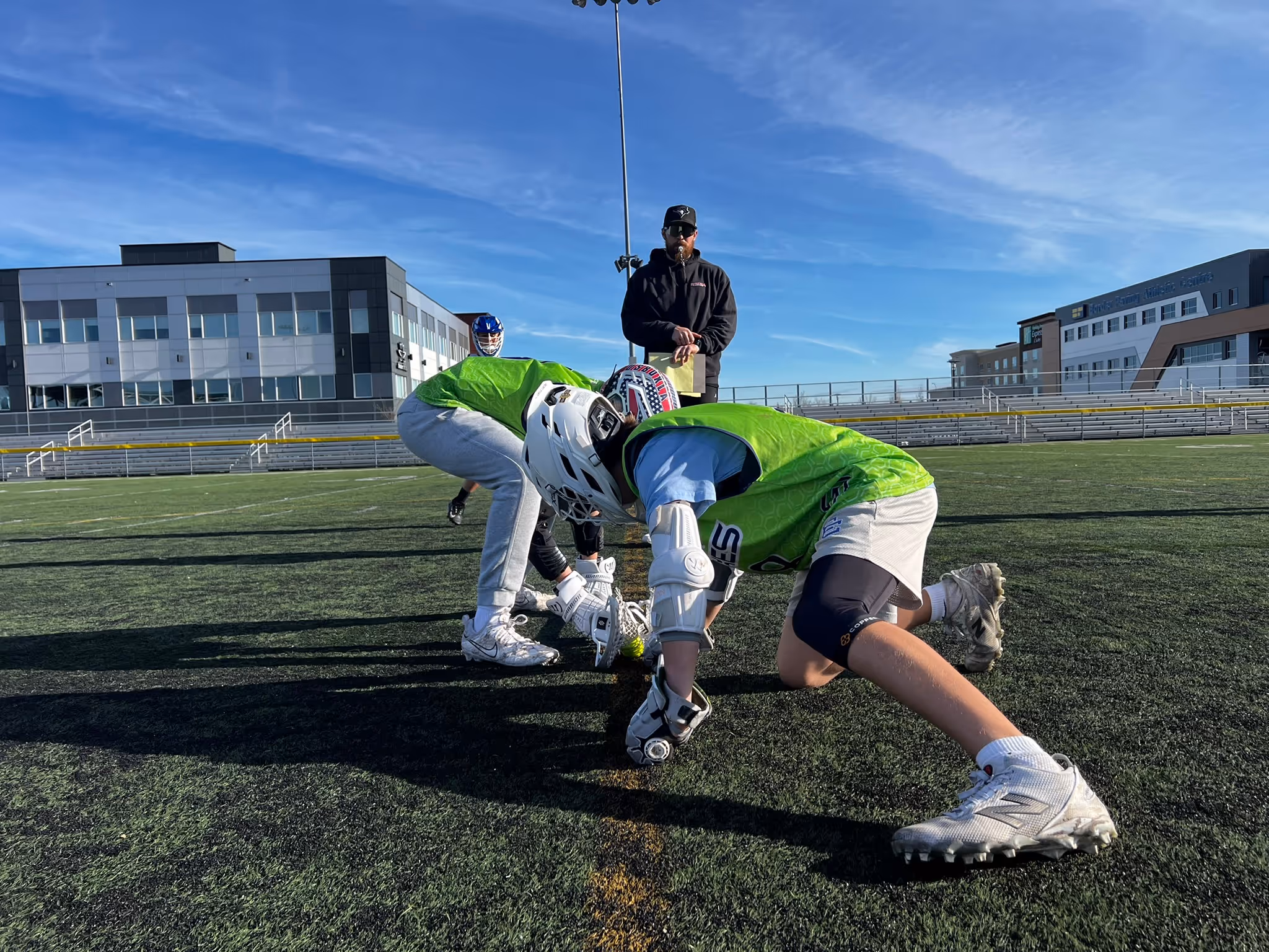 Two lacrosse players in green pinnies crouched face-off on a turf field with a coach standing behind them.