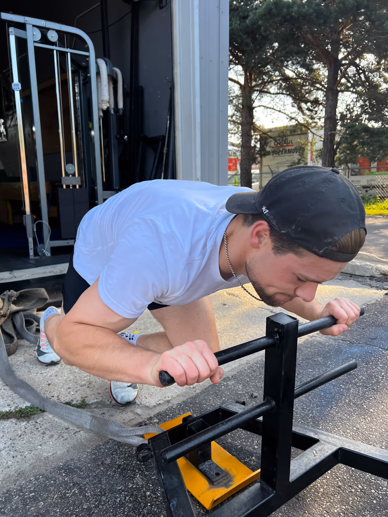 Man in white shirt and black cap pushing a weighted sled during an outdoor workout.