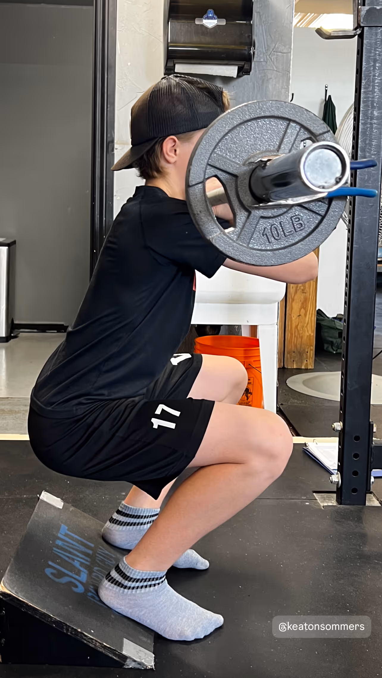 Youth in black clothes performing a barbell squat with 10lb weight plates on a ramp platform indoors.