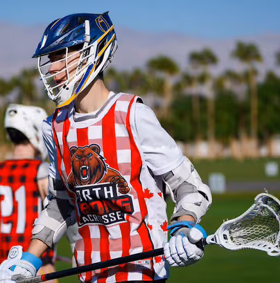 Youth lacrosse player in blue helmet and red striped jersey holding a lacrosse stick on a green field.