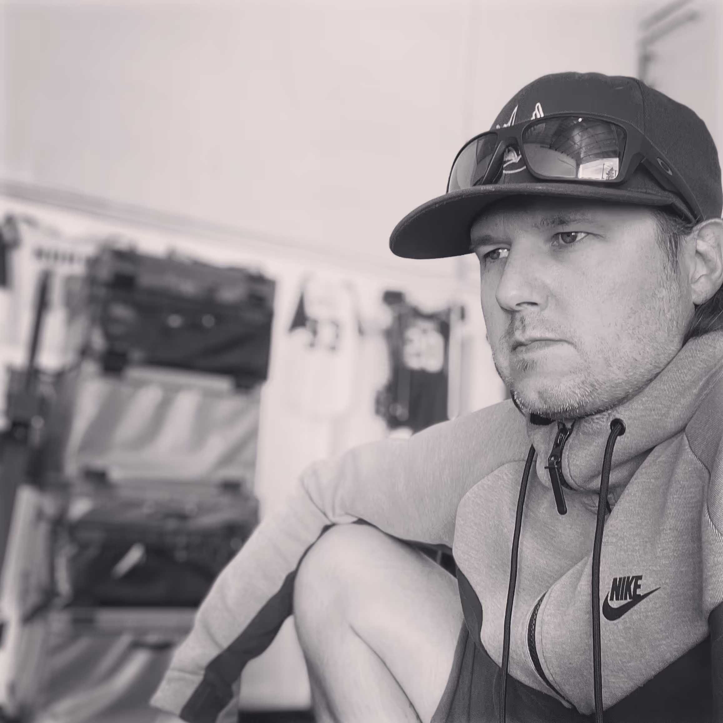 Man wearing a Nike hoodie, cap, and sunglasses resting with a thoughtful expression indoors.