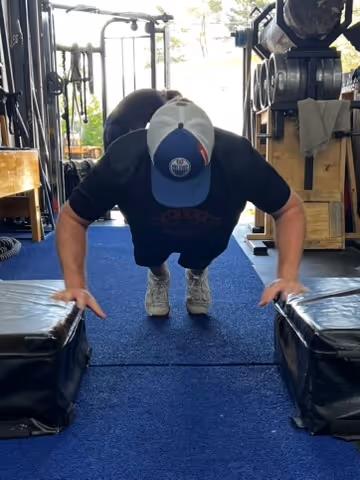Person wearing a cap performing elevated push-ups between two padded platforms in a gym.