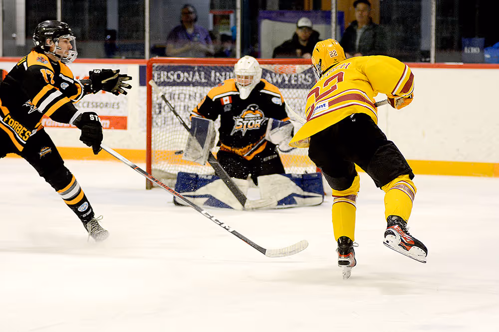 Hockey player in yellow jersey taking a shot on goal as goalie and defender in black jerseys prepare to block it.