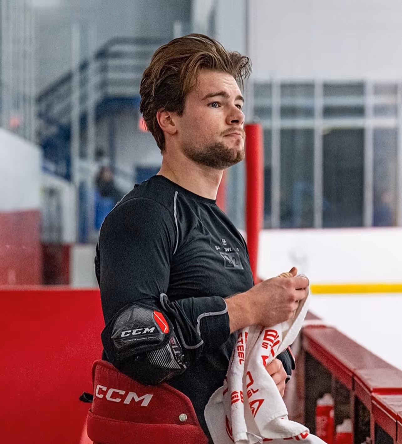 Hockey player wearing black protective gear and holding a white towel on the bench of an ice rink.