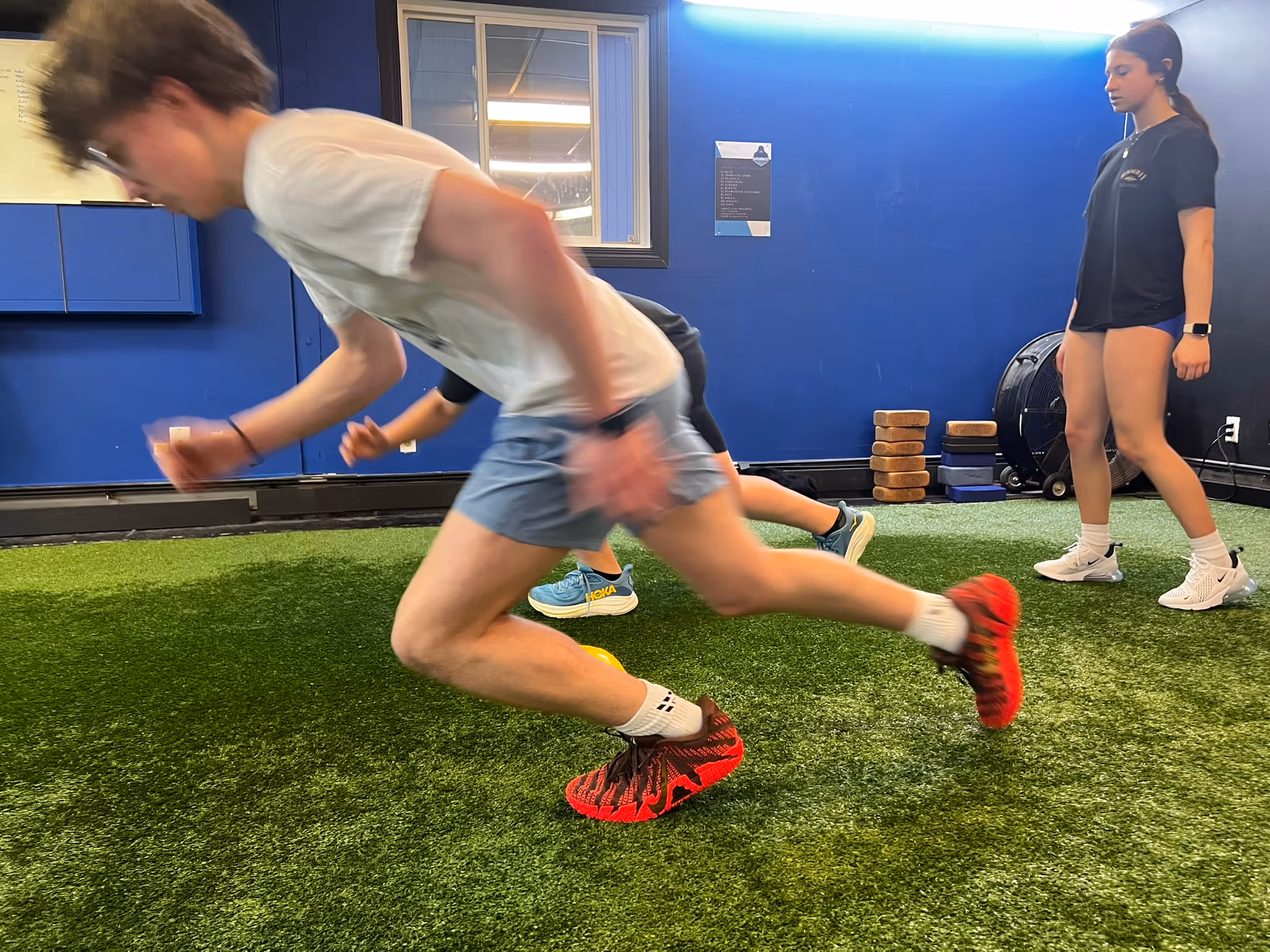 Three young people in a gym with artificial turf, two running and one standing watching.
