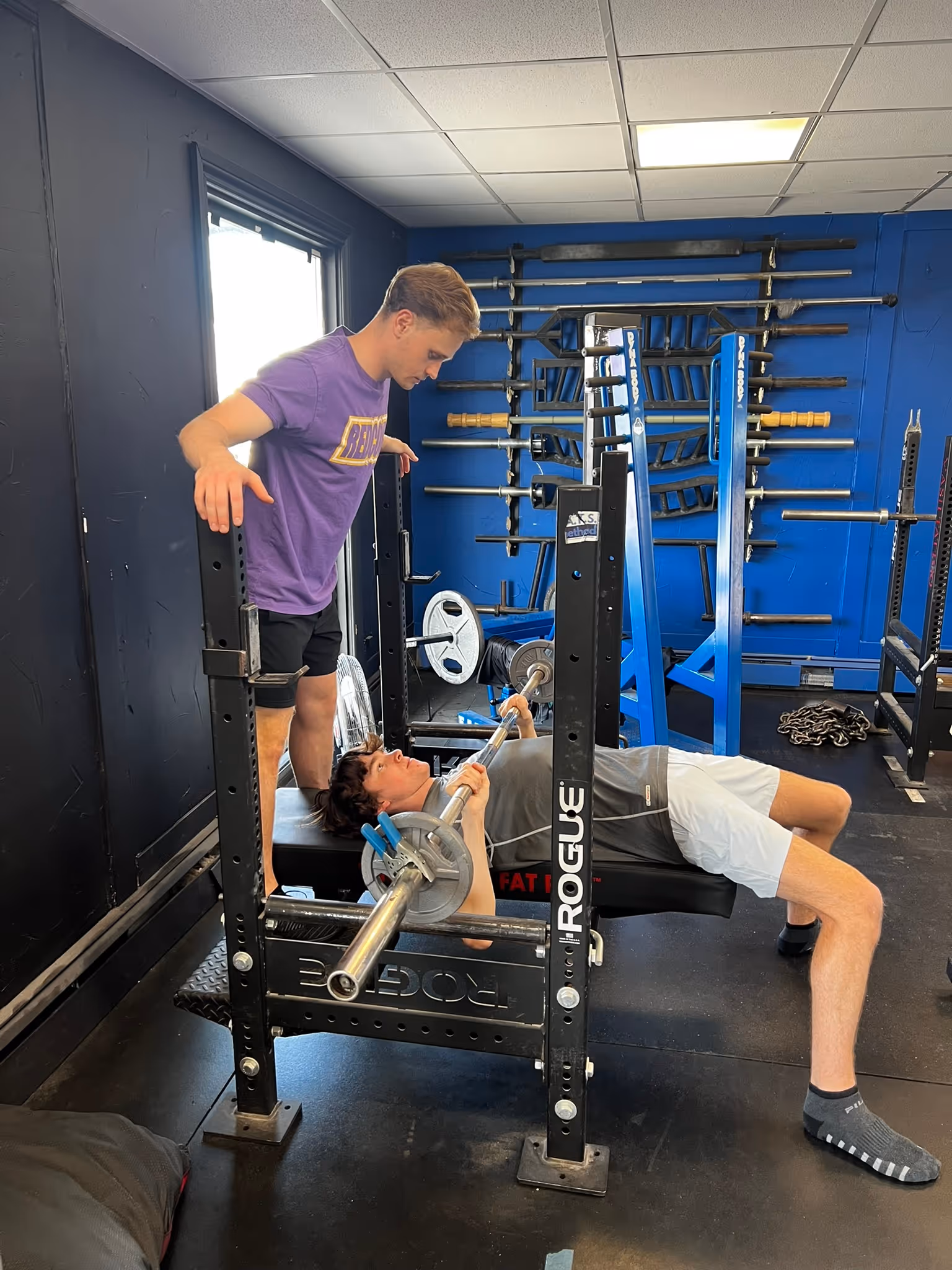 Man in purple shirt spotting another man lying on bench press lifting barbell with weights in gym.