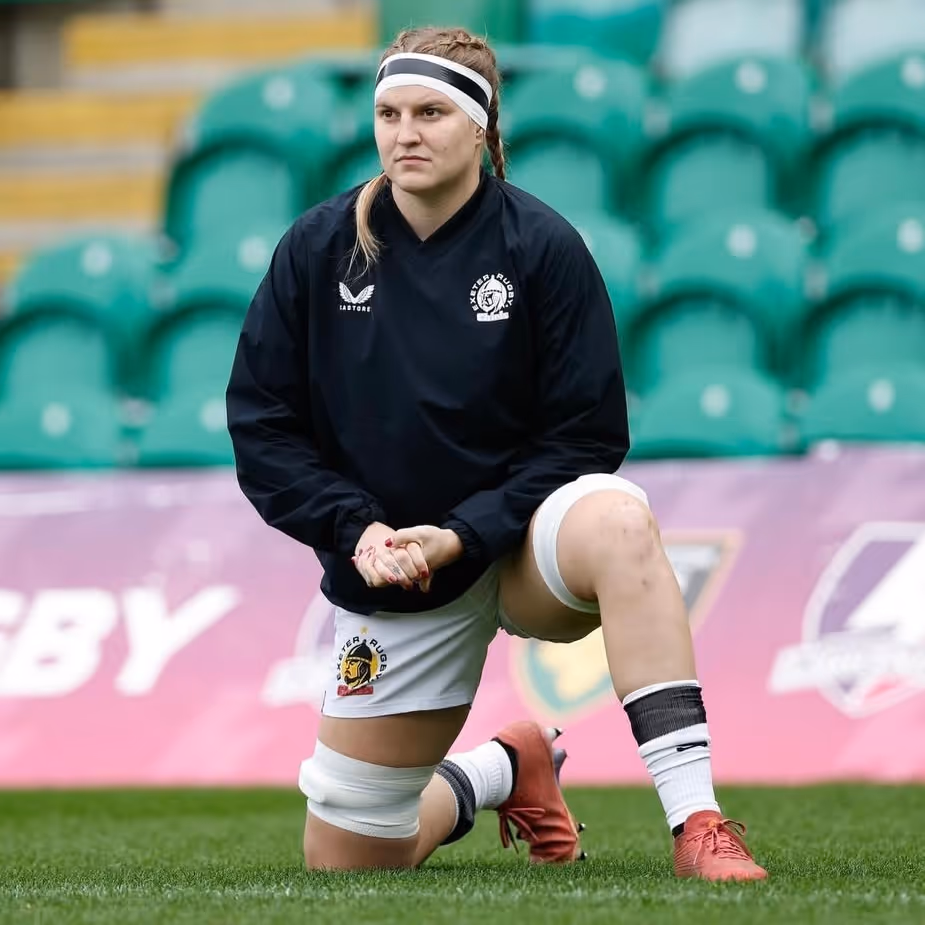 Female rugby player in black Exeter Rugby jacket stretching on grass field with green stadium seats behind.