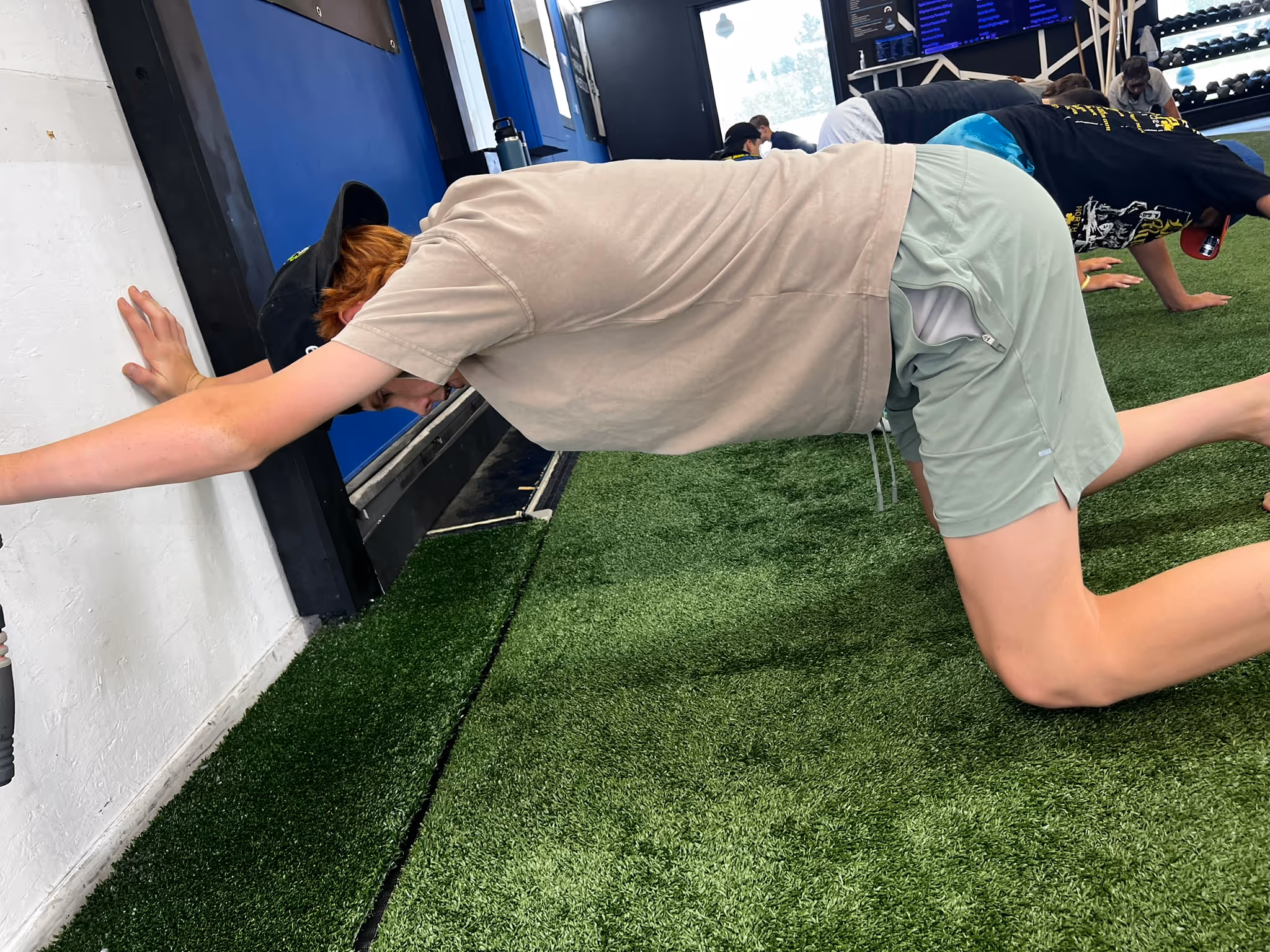 Person in beige shirt and green shorts stretching against a wall on artificial turf indoors.