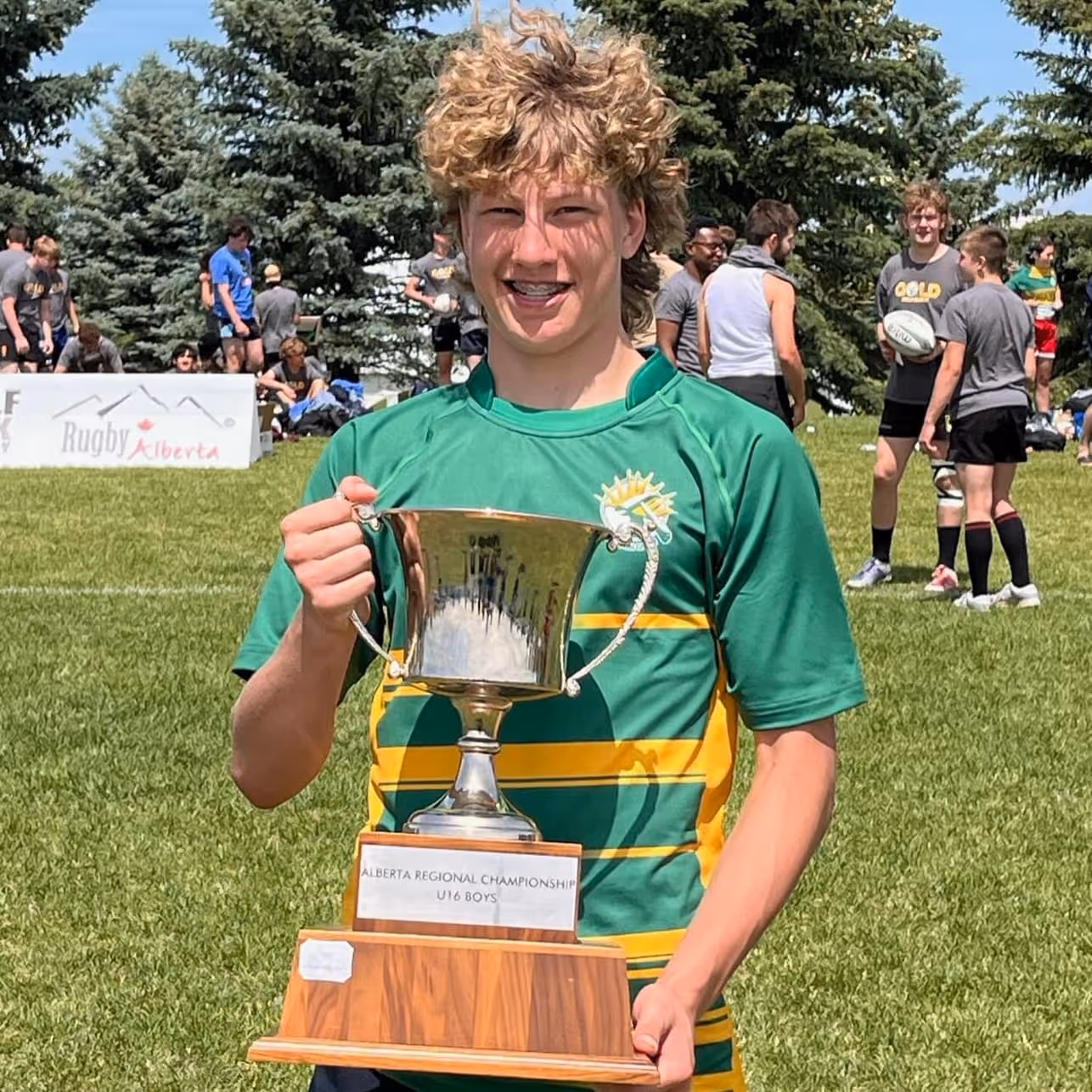 Teen rugby player in green and yellow holds Alberta Regional Championship U16 Boys trophy on field.