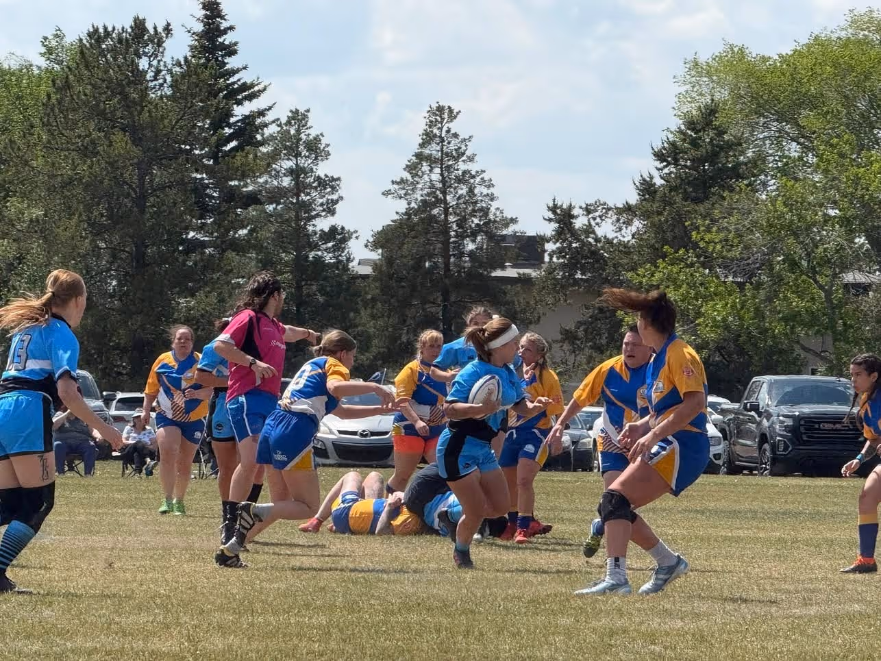 Women’s rugby players in blue and yellow uniforms compete on a grassy field with trees and parked cars in the background.