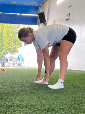 Woman in athletic wear bending forward to touch her toes on artificial turf indoors.