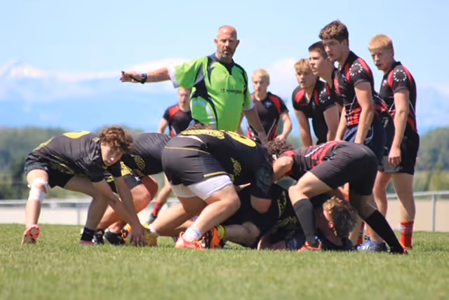 Youth rugby players in black and red uniforms engaged in a scrum on grass with a referee in a green shirt signaling.