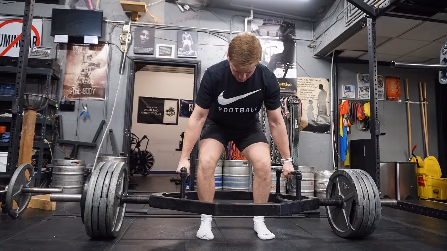 Man lifting a heavily weighted hex bar in a gym, preparing to deadlift.