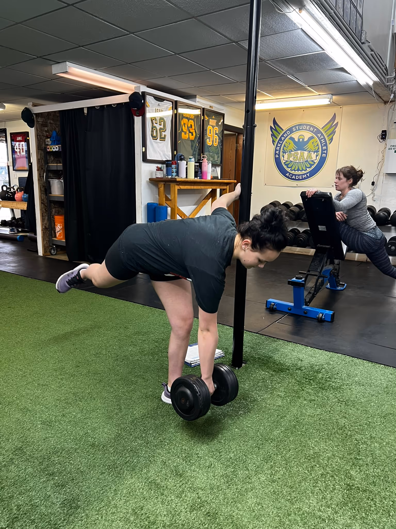 Two women exercising indoors, one performing a single-leg deadlift with dumbbells, the other stretching on a bench.
