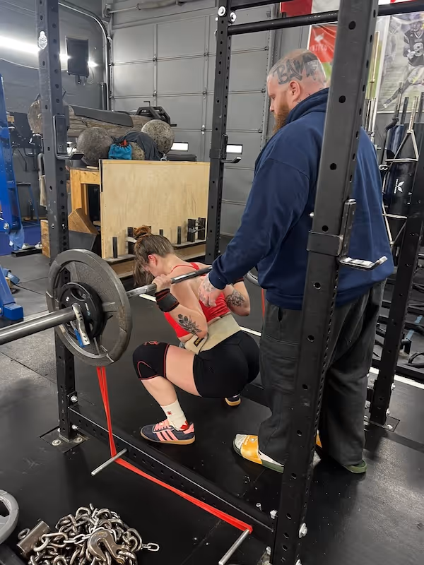 Woman with tattoos performing a barbell squat in a gym, assisted by a man standing behind her.