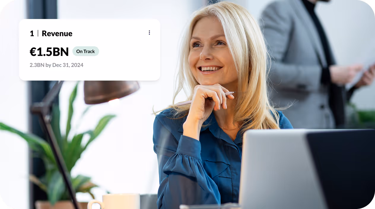 Smiling woman with blonde hair sitting at a desk with a laptop, with a plant and a man in the background, alongside a graphic showing revenue of €1.5BN marked as On Track.