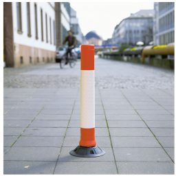 Orange and white traffic bollard on a paved sidewalk with a cyclist in the background.