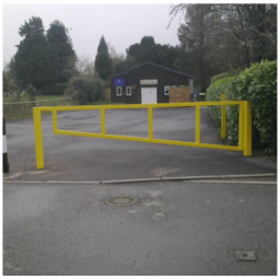 Yellow metal barrier gate blocking a paved driveway leading to a small brick building surrounded by trees and bushes.