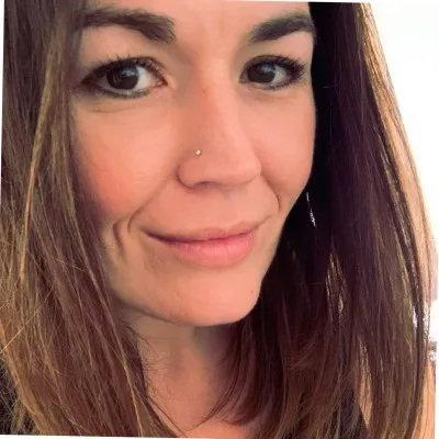 Close-up of a woman with long brown hair, brown eyes, and a nose piercing, smiling slightly.