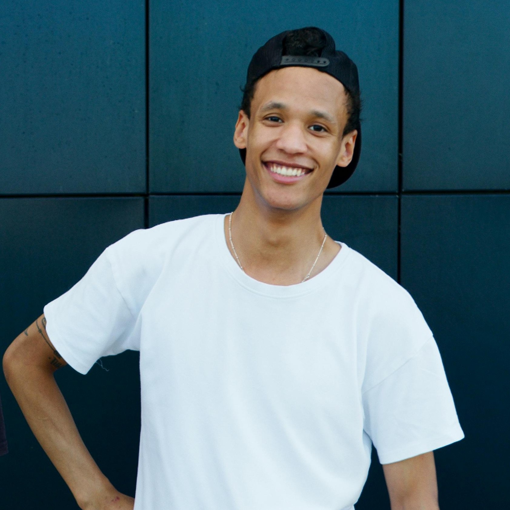 Smiling young man wearing a white t-shirt and black backward cap standing against a dark tiled wall.