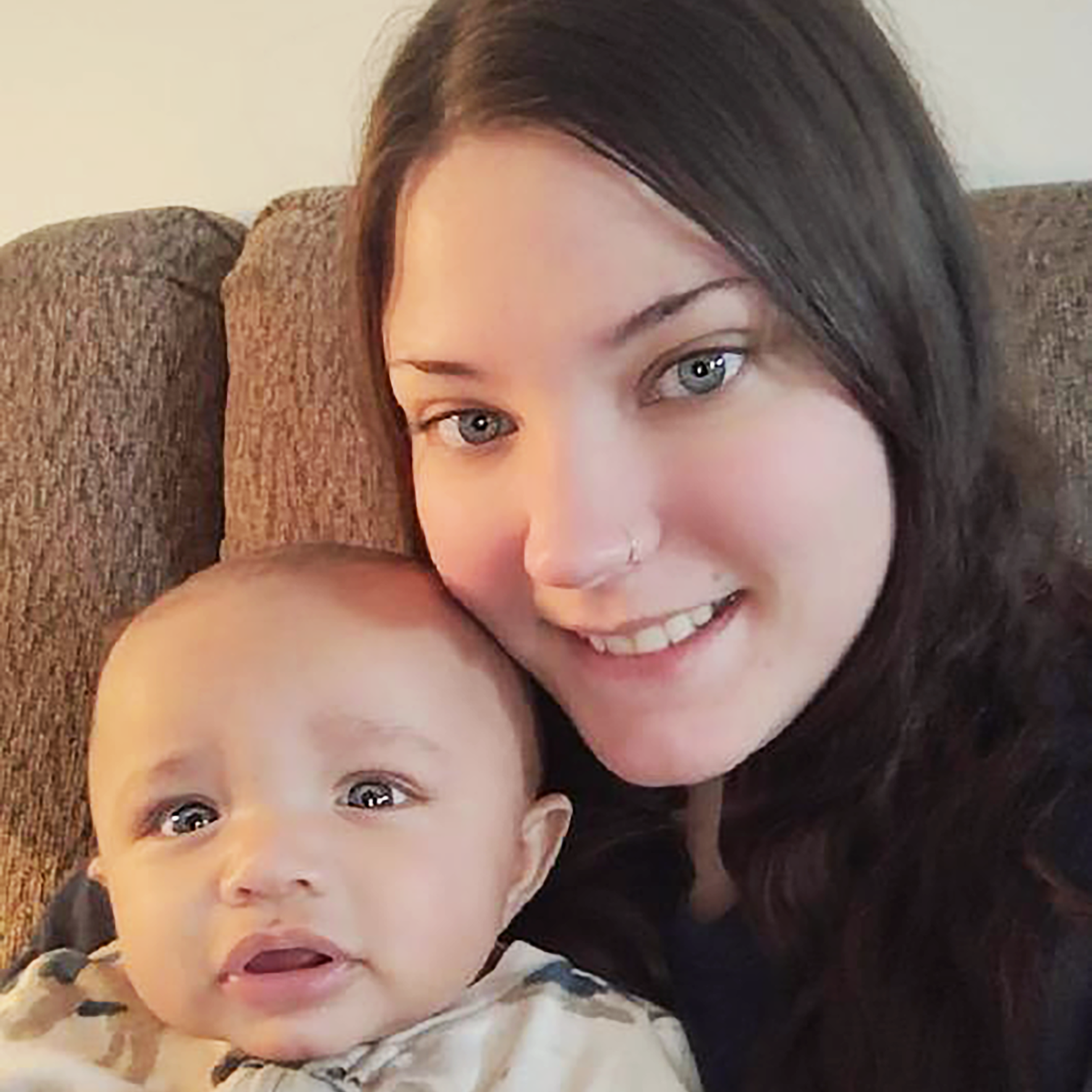 Woman with long dark hair and blue eyes smiling next to a baby with light brown skin and big eyes on a brown couch.
