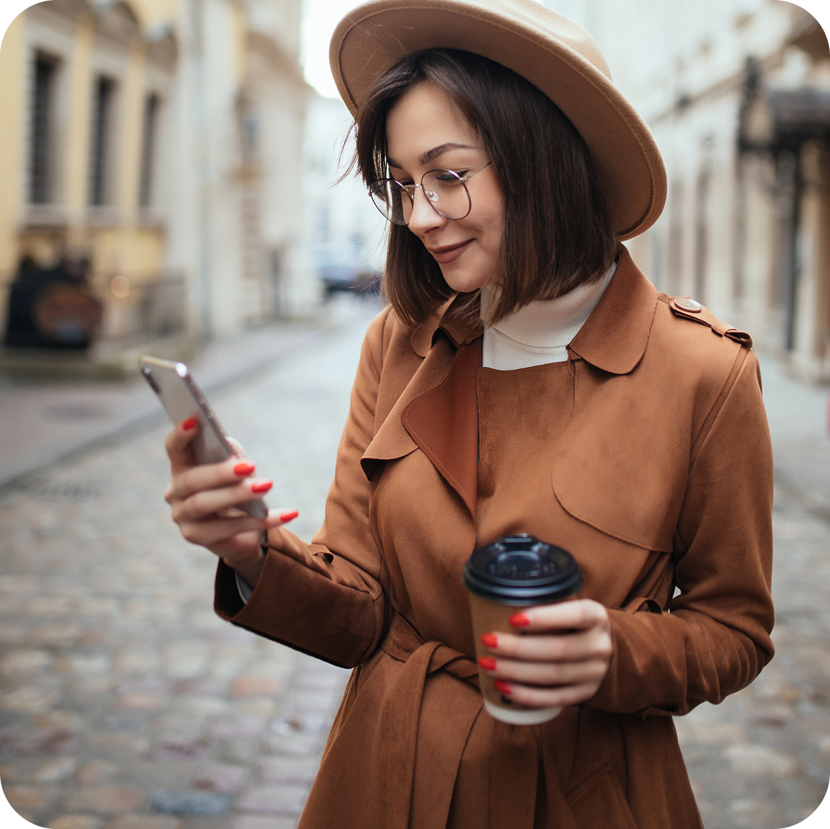 Woman in a brown coat checking her phone while holding a coffee