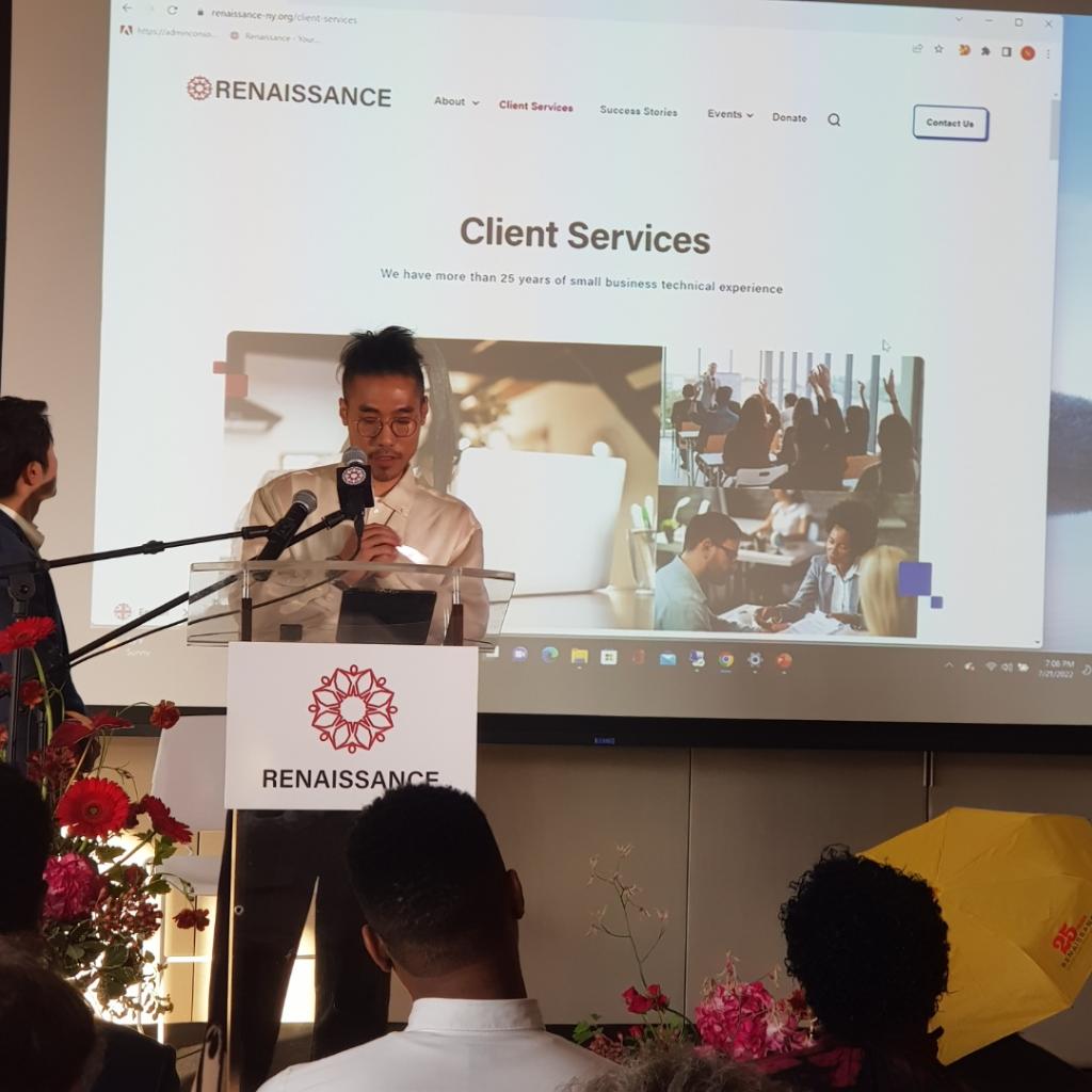 Man wearing glasses speaking at a podium with a Renaissance sign in front, delivering a presentation with a projected webpage titled Client Services behind him.