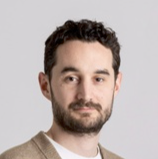 Portrait of a man with dark curly hair, beard, and mustache wearing a beige sweater.