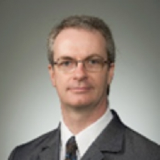 Portrait of a man with glasses wearing a dark suit, white shirt, and patterned tie against a neutral background.
