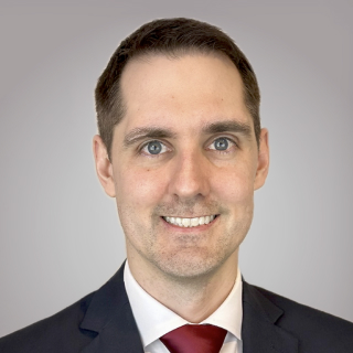 Smiling man with short dark hair wearing a dark suit, white shirt, and red tie against a gray background.