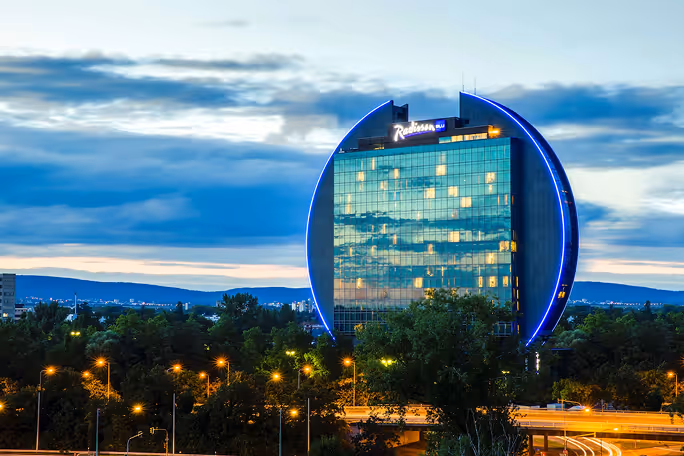 Radisson Blu hotel building lit with blue lines at dusk, surrounded by trees and city lights.