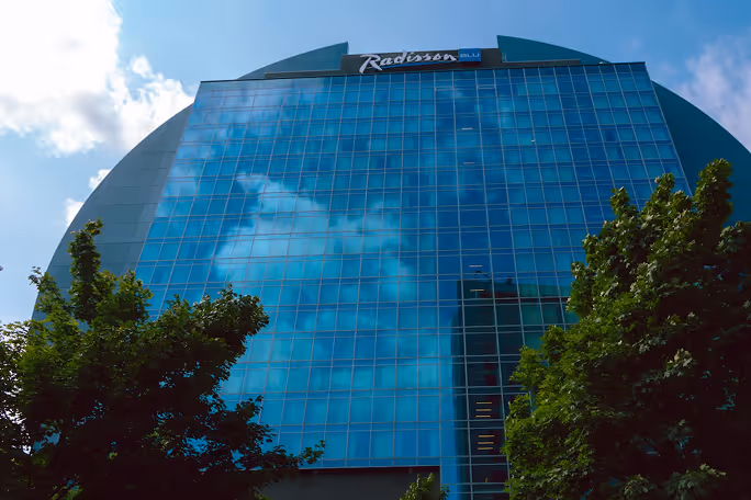 Glass facade of Radisson Blu hotel reflecting clouds with green trees at the bottom.