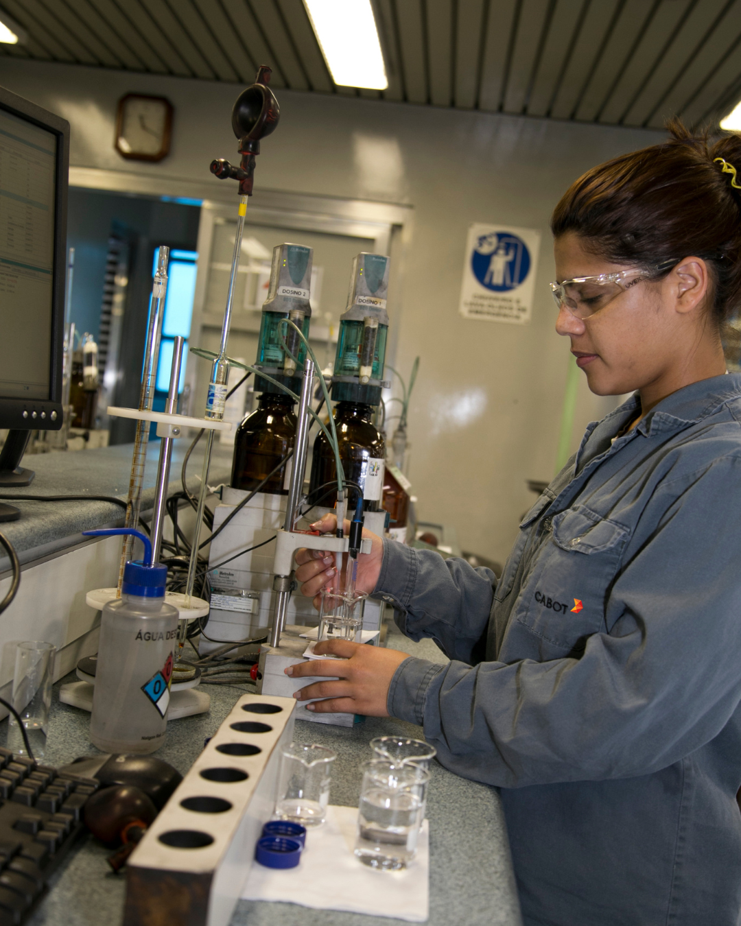 A Cabot lab technician in safety glasses runs a titration at a quality control bench with automated dosing units and glassware, beside a monitor of test data.