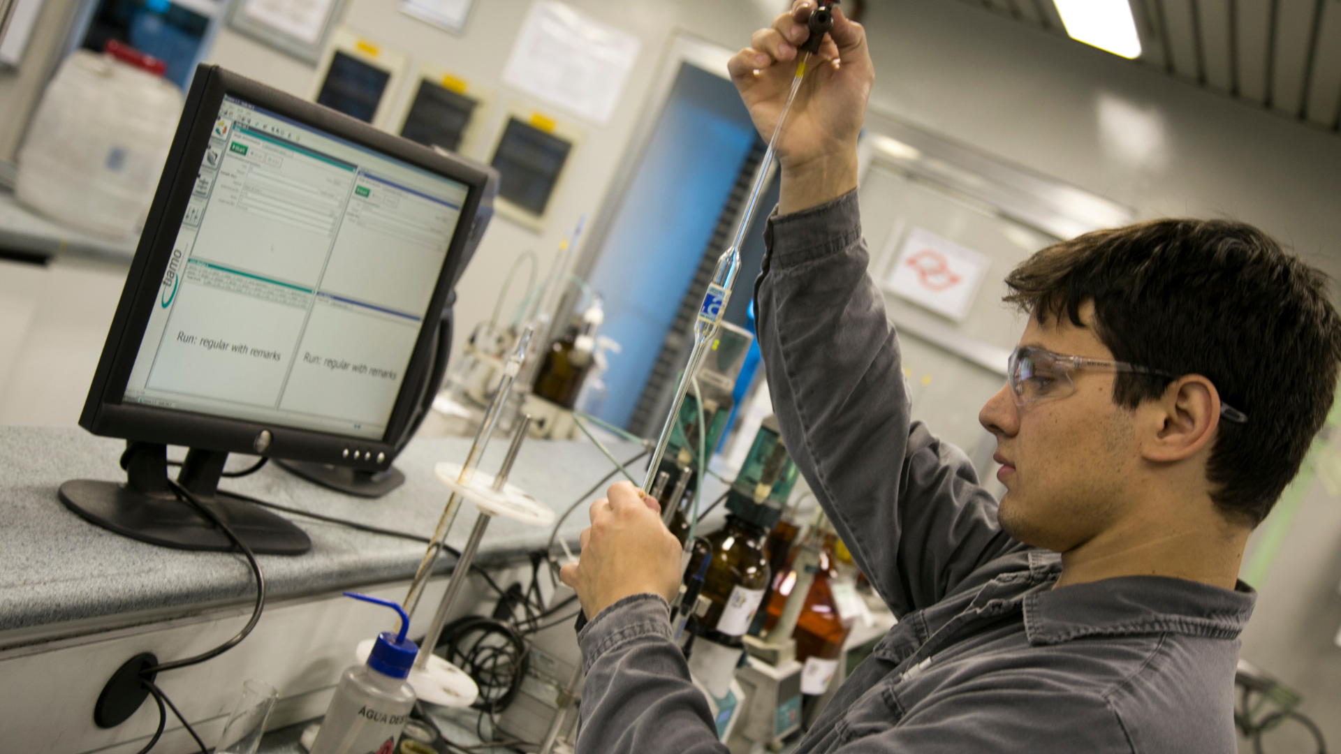 A young lab technician in safety glasses holds up a glass pipette to check a measurement, next to a computer showing quality control test records and reagent bottles on the bench.