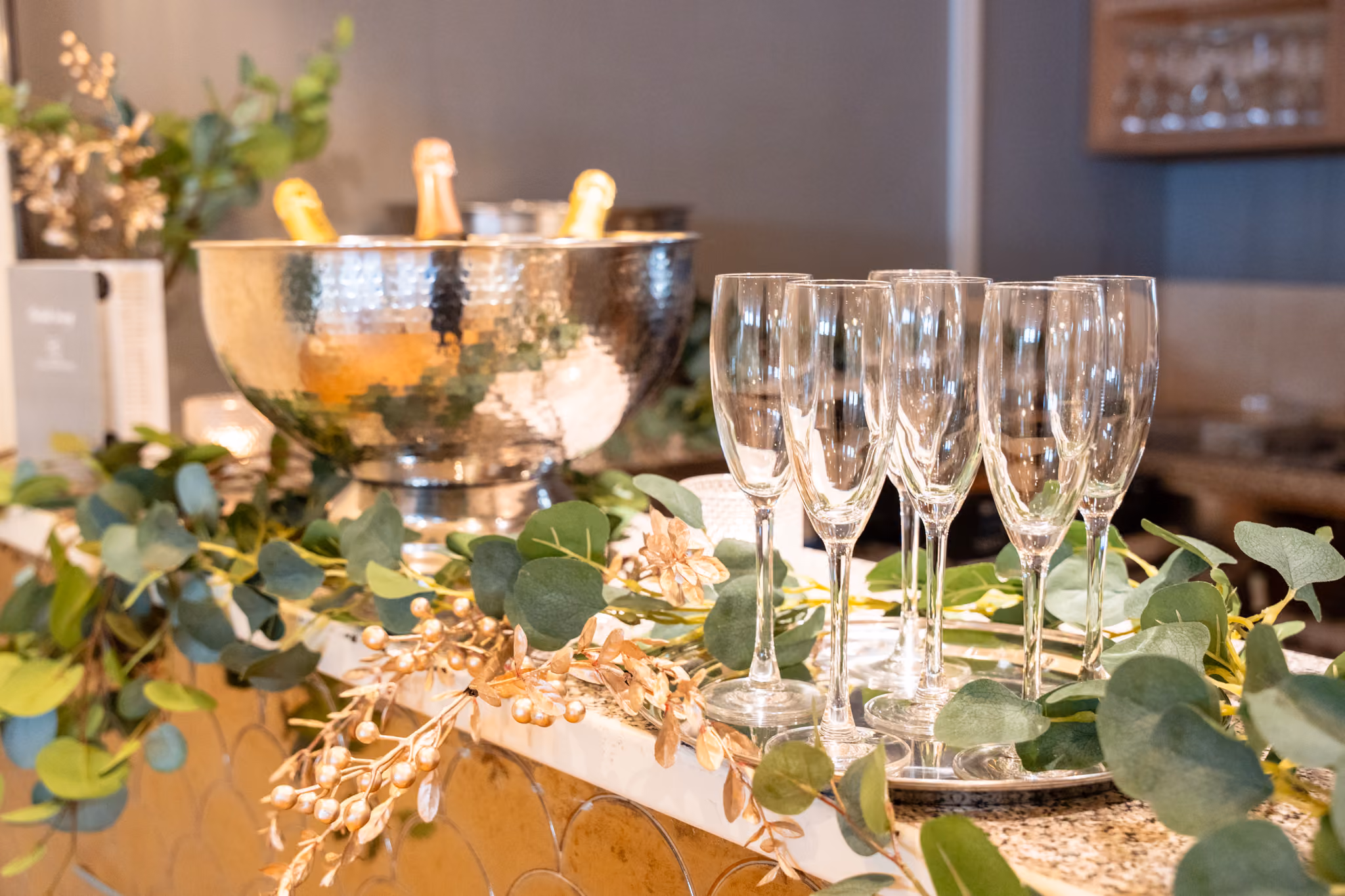 A wedding drinks station styled with eucalyptus garland and gold botanical accents. Five empty champagne flutes stand on a silver tray in the foreground, with a large hammered silver ice bucket holding two champagne bottles in the background.