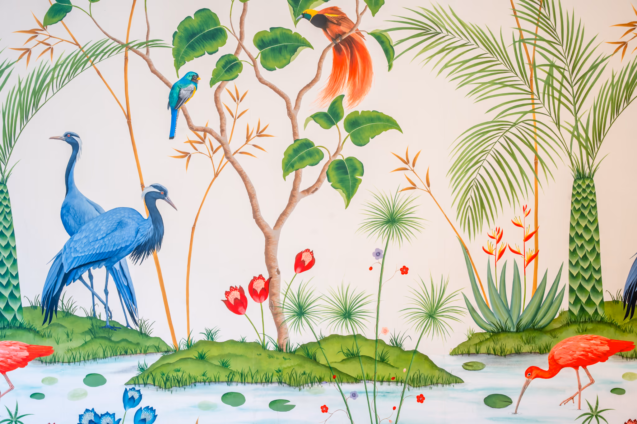 A detailed botanical mural featuring exotic birds and tropical foliage. Two blue herons stand on a grassy island beside a pond with lily pads, while red ibises wade in the foreground. A bird of paradise with vivid orange plumage perches in a tree above a small teal kingfisher. Palm fronds, banana leaves, bamboo, red tulips, and heliconia flowers complete the scene against a cream background.