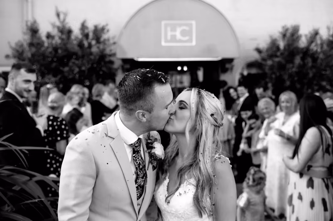 A bride and groom share a kiss outside their wedding venue, confetti scattered in their hair and on their clothing. The groom wears a light suit with a floral tie and buttonhole, the bride in a lace dress with her blonde hair half up. Wedding guests celebrate in the background outside a venue entrance marked with an HC logo.