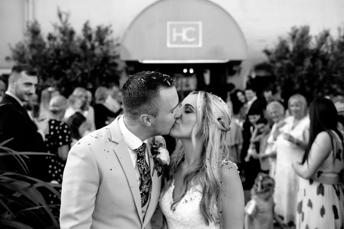 A bride and groom share a kiss outside their wedding venue, confetti scattered in their hair and on their clothing. The groom wears a light suit with a floral tie and buttonhole, the bride in a lace dress with her blonde hair half up. Wedding guests celebrate in the background outside a venue entrance marked with an HC logo.