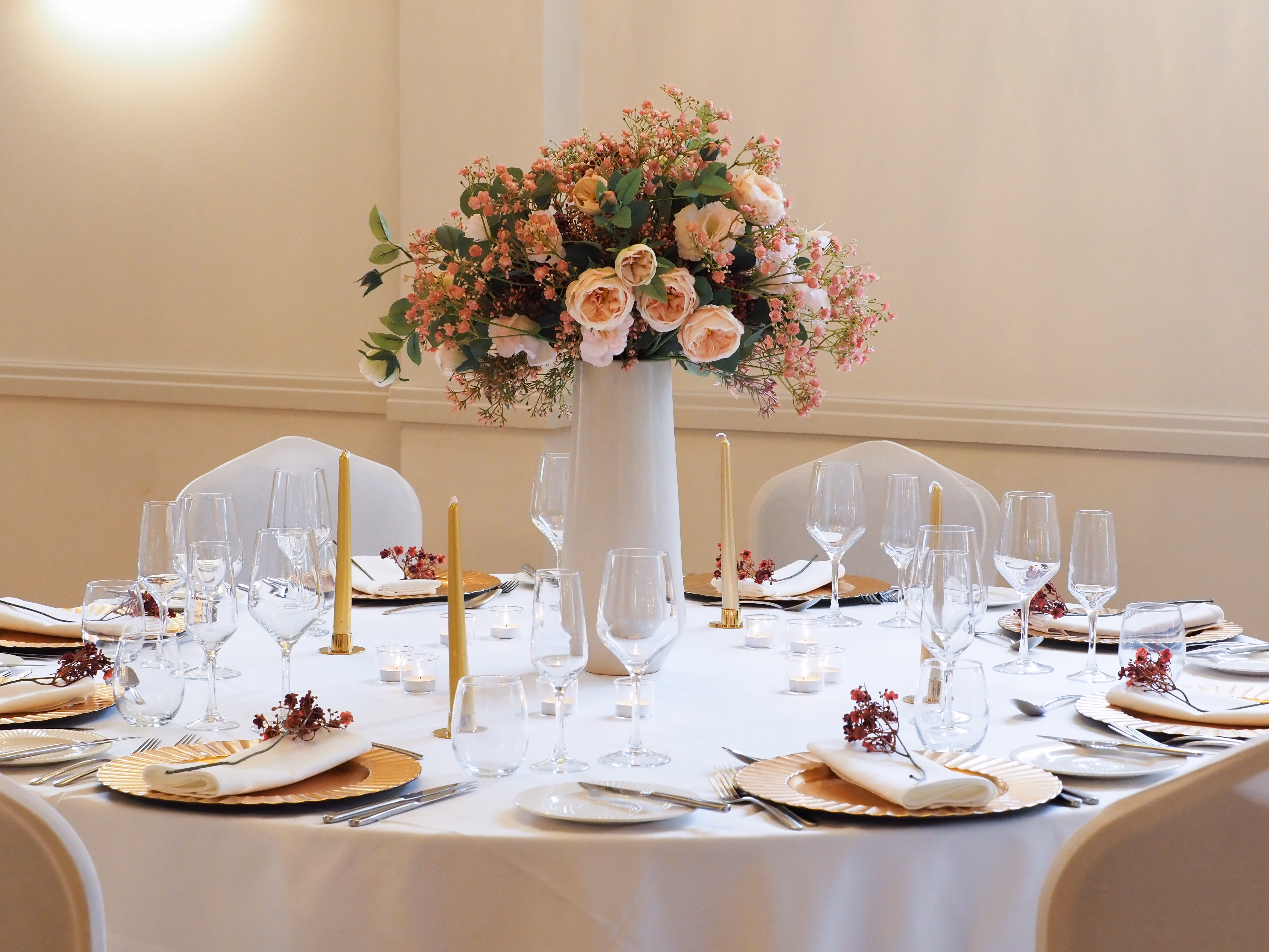 An elegantly dressed round wedding reception table with a white linen cloth. A tall white vase centrepiece holds a full arrangement of blush roses, pink gypsophila, and eucalyptus. Place settings feature gold charger plates, white folded napkins with small dried flower details, multiple wine glasses, and slim gold taper candles with tealights scattered between settings.