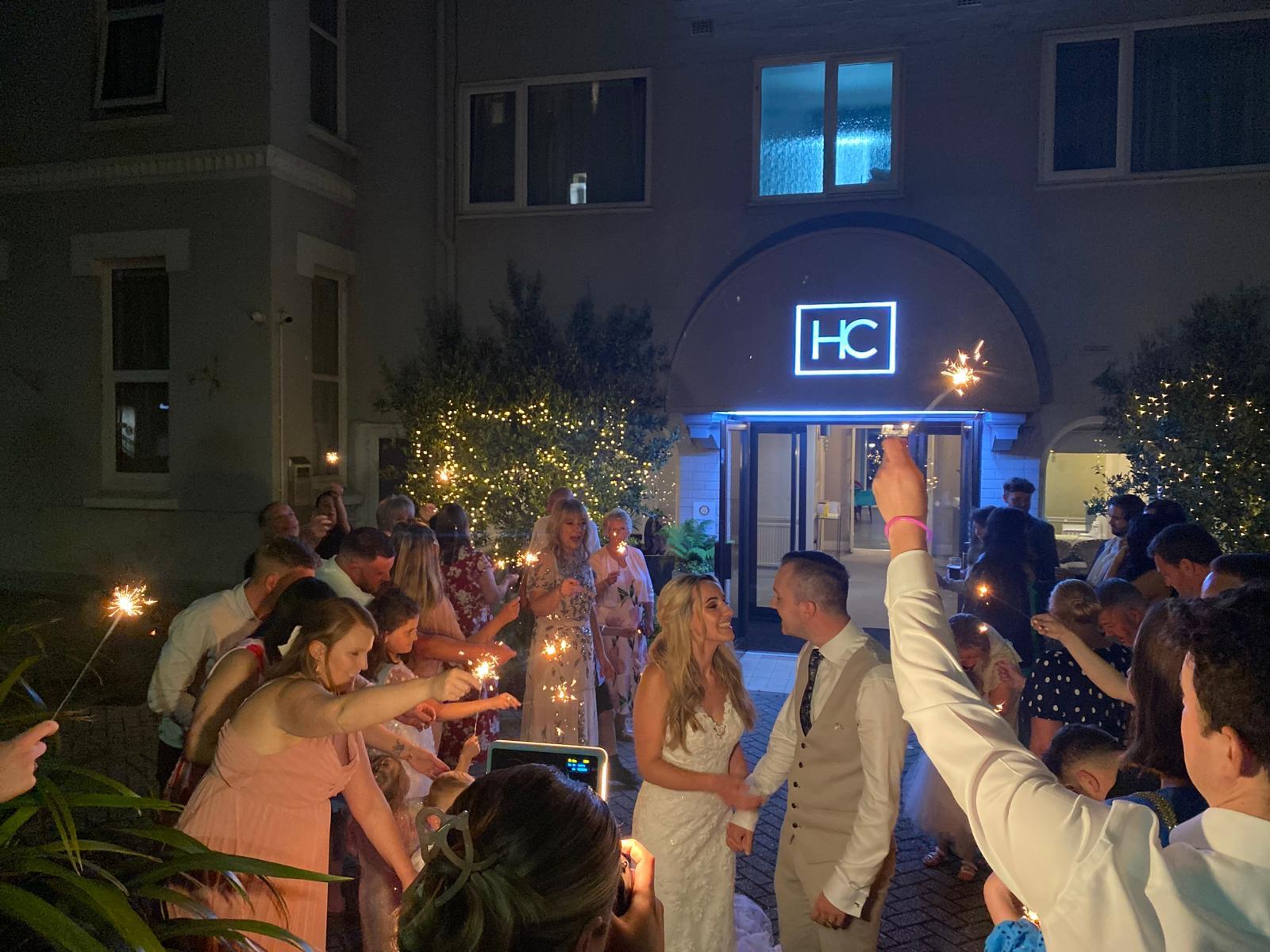 A bride and groom walk through a sparkler send-off outside Hotel Collingwood at night, surrounded by cheering wedding guests holding lit sparklers. The bride wears a lace dress and smiles broadly as the groom, in a light suit and waistcoat, raises a sparkler above the crowd. The hotel's illuminated HC sign glows blue above the arched entrance behind them.