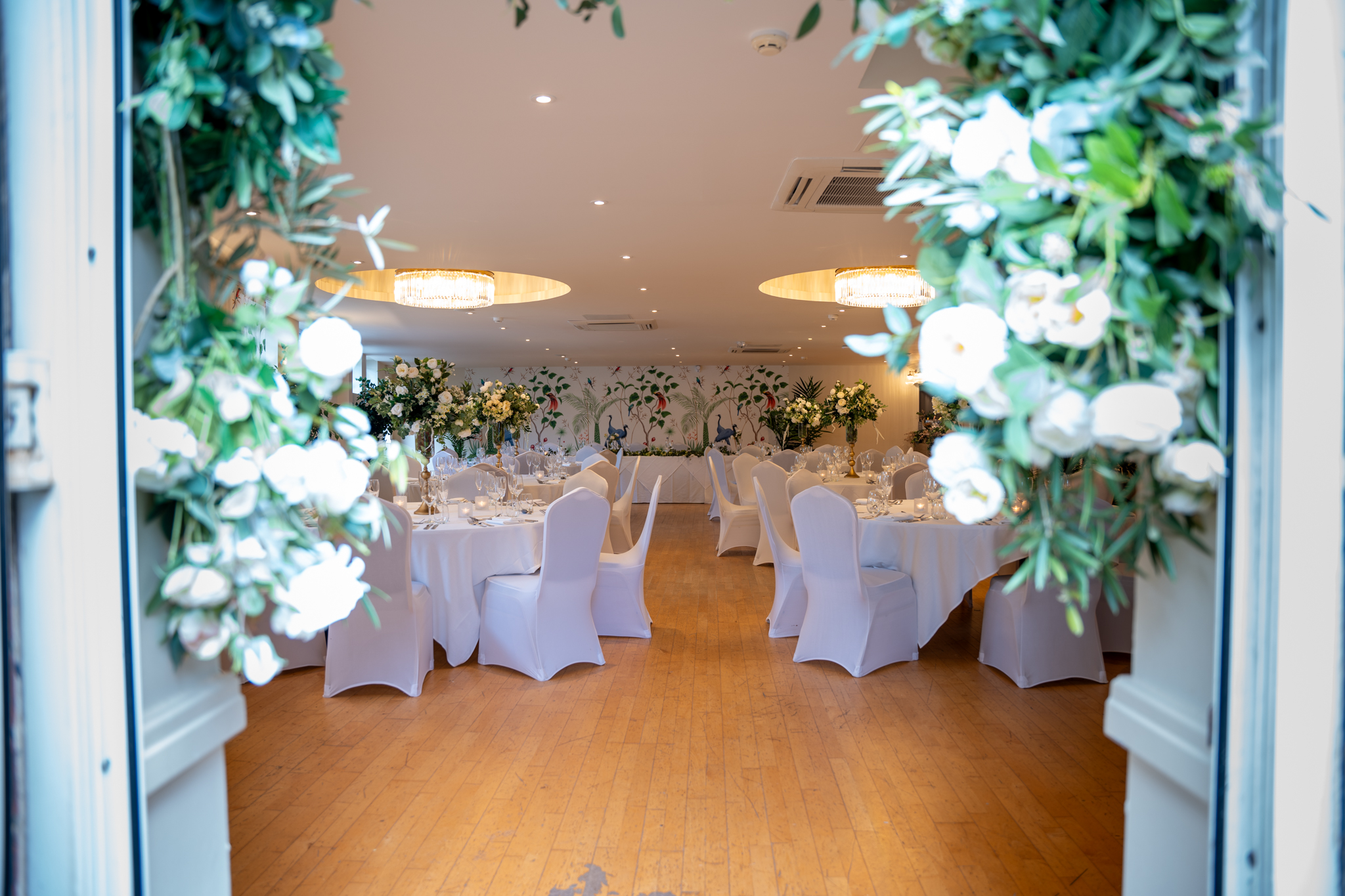 A wedding reception room viewed through open doors framed with lush greenery and white flowers. Inside, round tables dressed in white linen with white chair covers are arranged on a warm wooden floor beneath two large gold circular chandeliers. Tall floral arrangements in gold vases line the top table at the far end, set against the room's distinctive botanical bird mural.