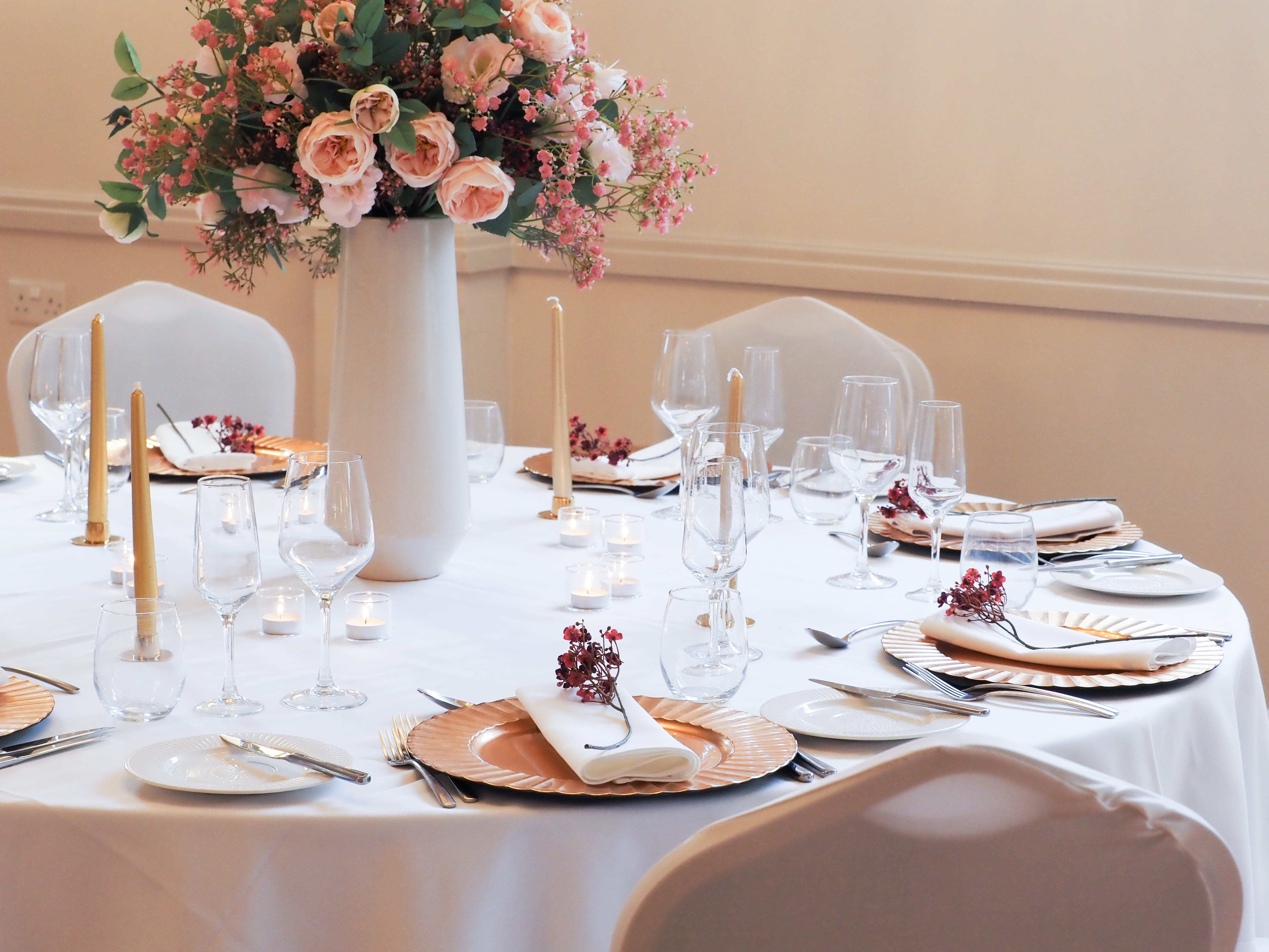 A close-up of a dressed wedding reception table with a white linen cloth. Gold charger plates hold white folded napkins tied with small dried dark red flower sprigs. Multiple wine and champagne glasses are set at each place alongside silver cutlery. Slim gold taper candles and lit tealights are dotted around the table, with a tall white vase of blush roses and pink gypsophila as the centrepiece.