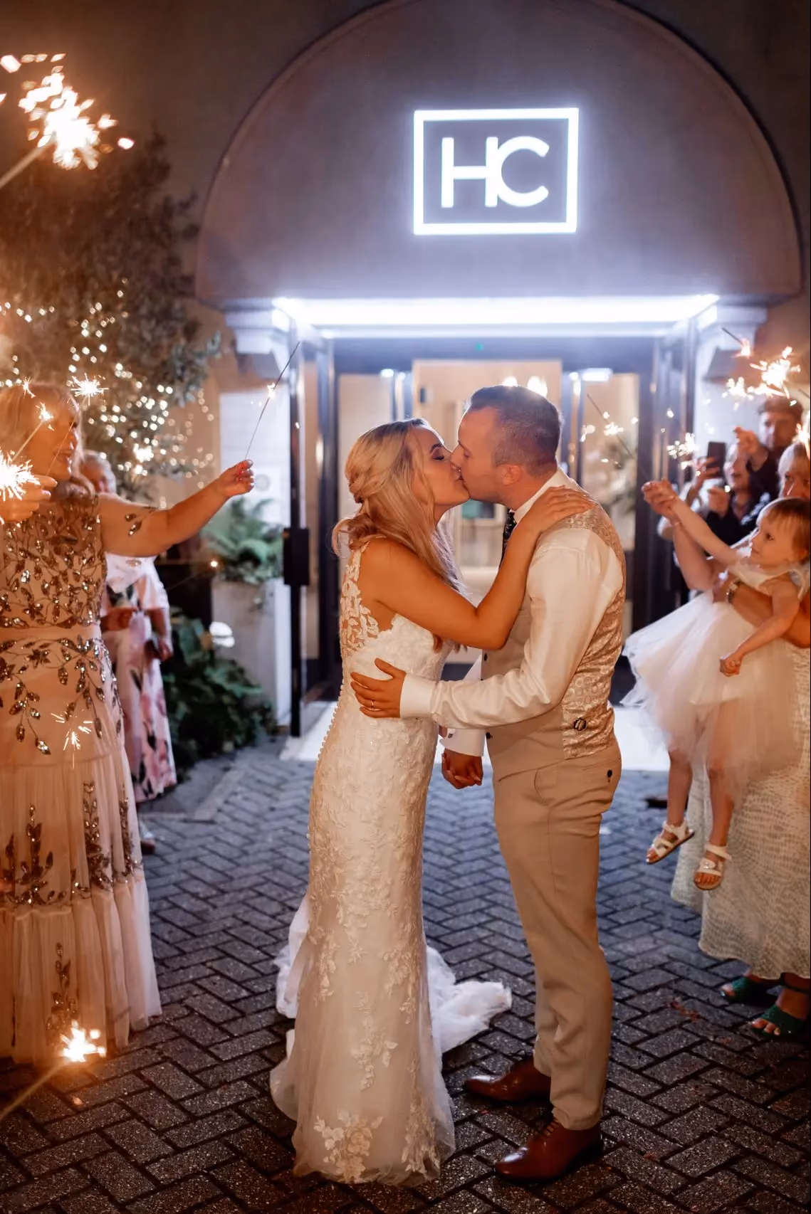 A bride and groom share a kiss during their sparkler exit outside Hotel Collingwood at night. The bride wears a fitted lace wedding dress with a train, the groom a cream waistcoat and trousers. Guests line either side holding lit sparklers, with a young flower girl in a white tutu visible to the right. The hotel's illuminated HC sign glows above the arched entrance behind them.
