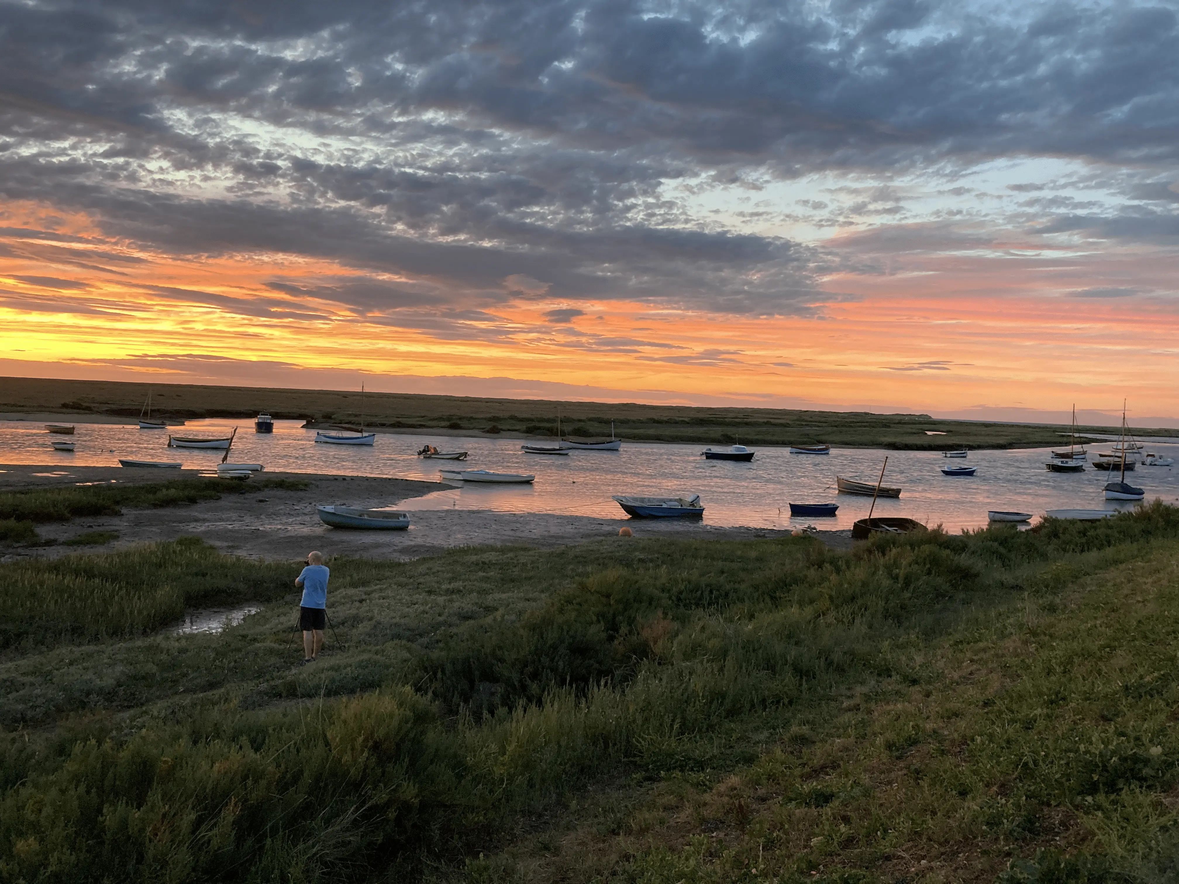 Boats anchored in marshy inlet during dramatic orange and gray sunset