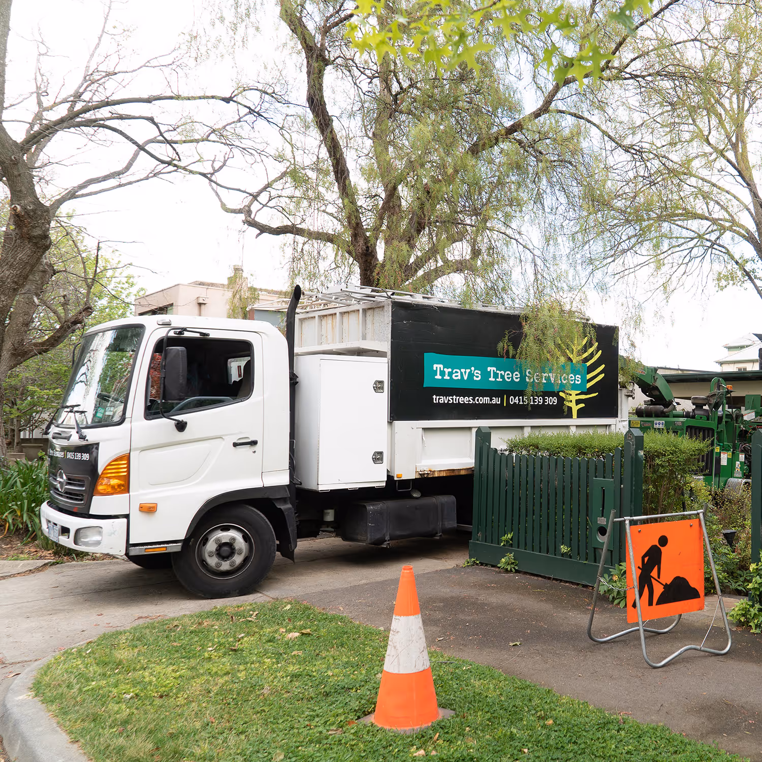 Trav's Tree Services white Hino truck parked on a Melbourne street, with a wood chipper and orange safety cone nearby.