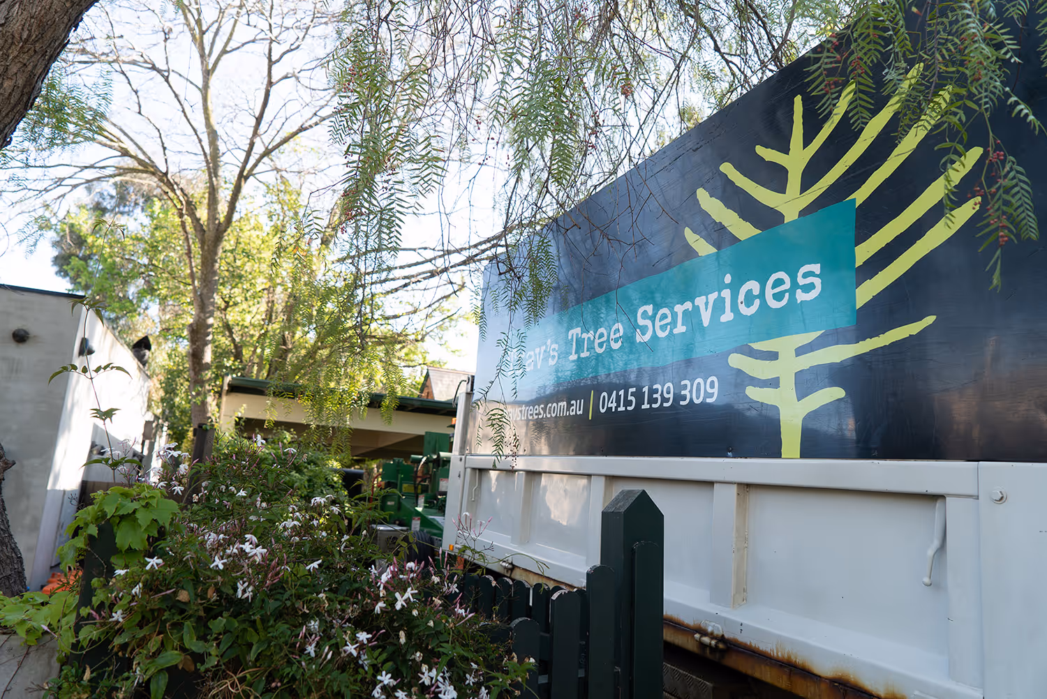 A Trav's Tree Services truck side panel showing the website and phone number, framed by green foliage and garden jasmine.