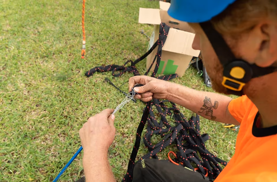 Worker in safety gear closely weaving rope strands during tree rigging preparation, surrounded by ropes and equipment.