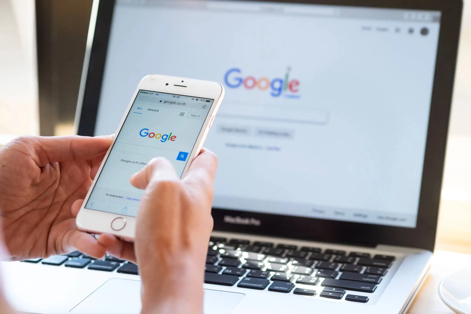 A person holds a smartphone displaying Google’s homepage while a laptop with the same interface is in the background