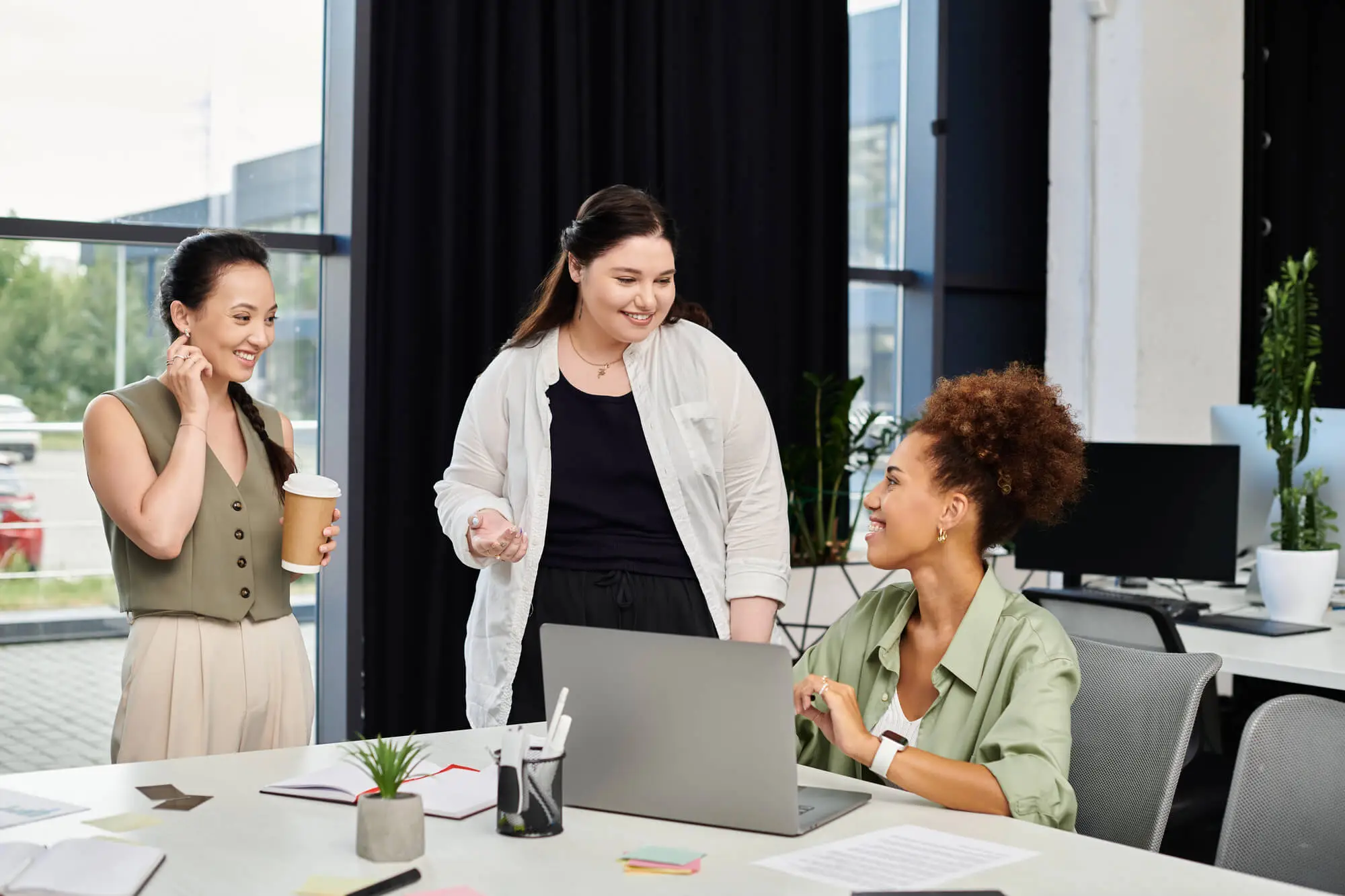 Three women engage in a discussion at a modern office setting, with a laptop and various office supplies on the table