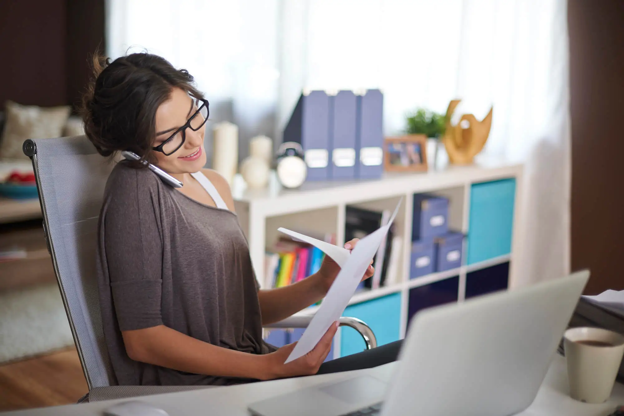 Person sitting in office chair, holding papers and talking on phone, with laptop and coffee cup on desk in bright workspace
